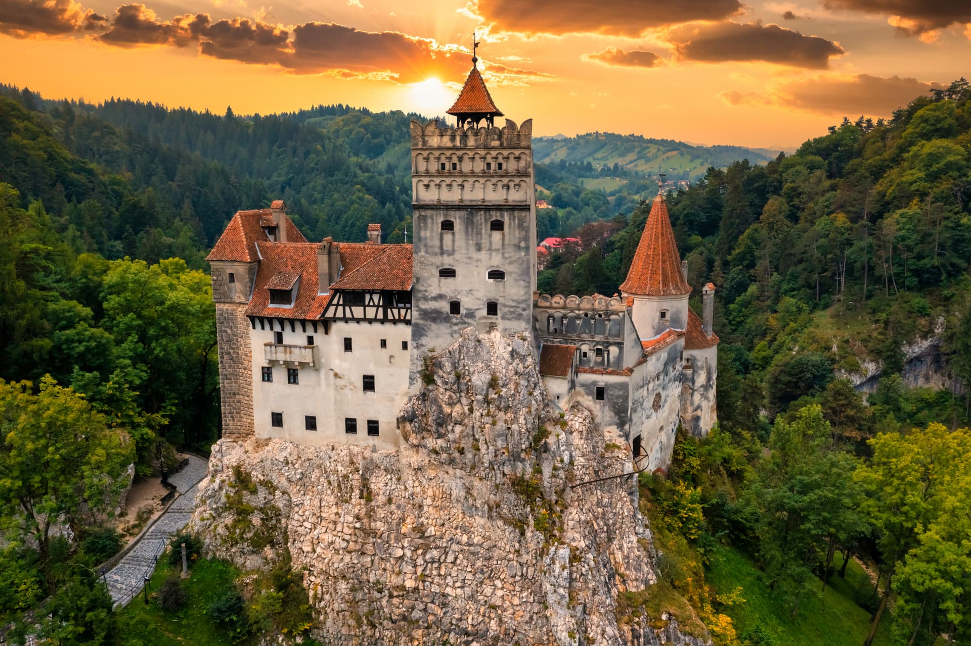Bran Castle in Transylvania, Romania, perched on a rocky outcrop at sunset over forested hills.