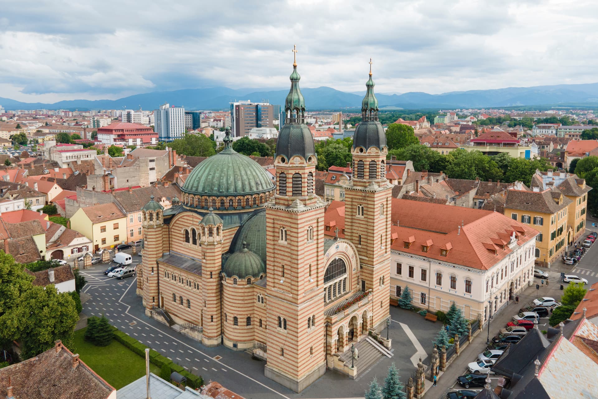 Cathedral of the Holy Trinity in Sibiu, Romania, with striped facade and green domes, overlooking city and mountains.