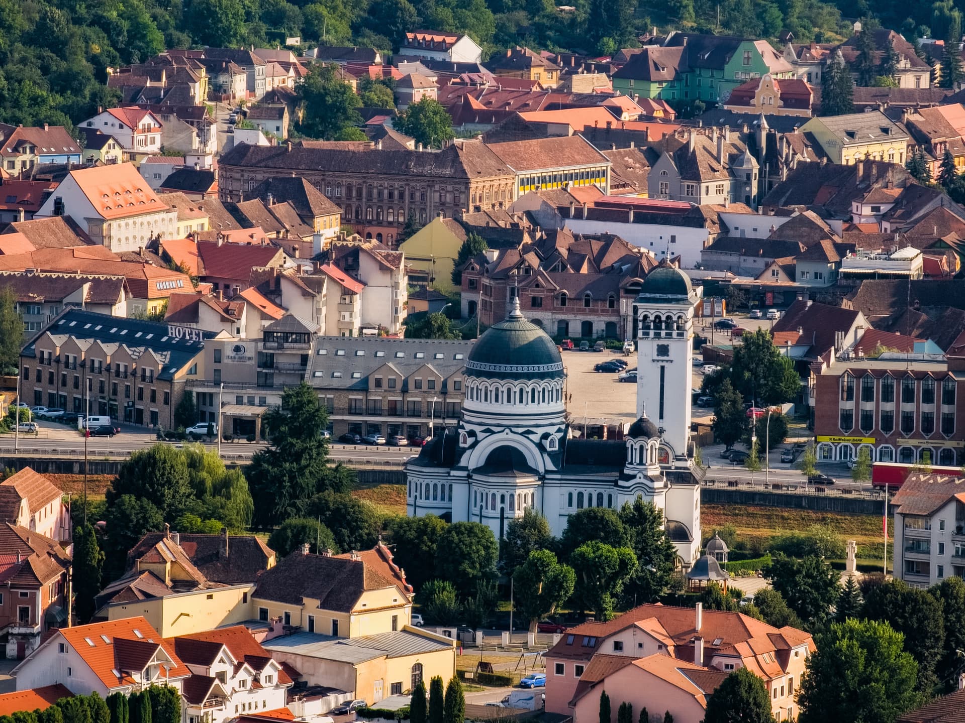 White Orthodox cathedral with green domes amidst dense housing and green hills in Sighisoara, Romania.