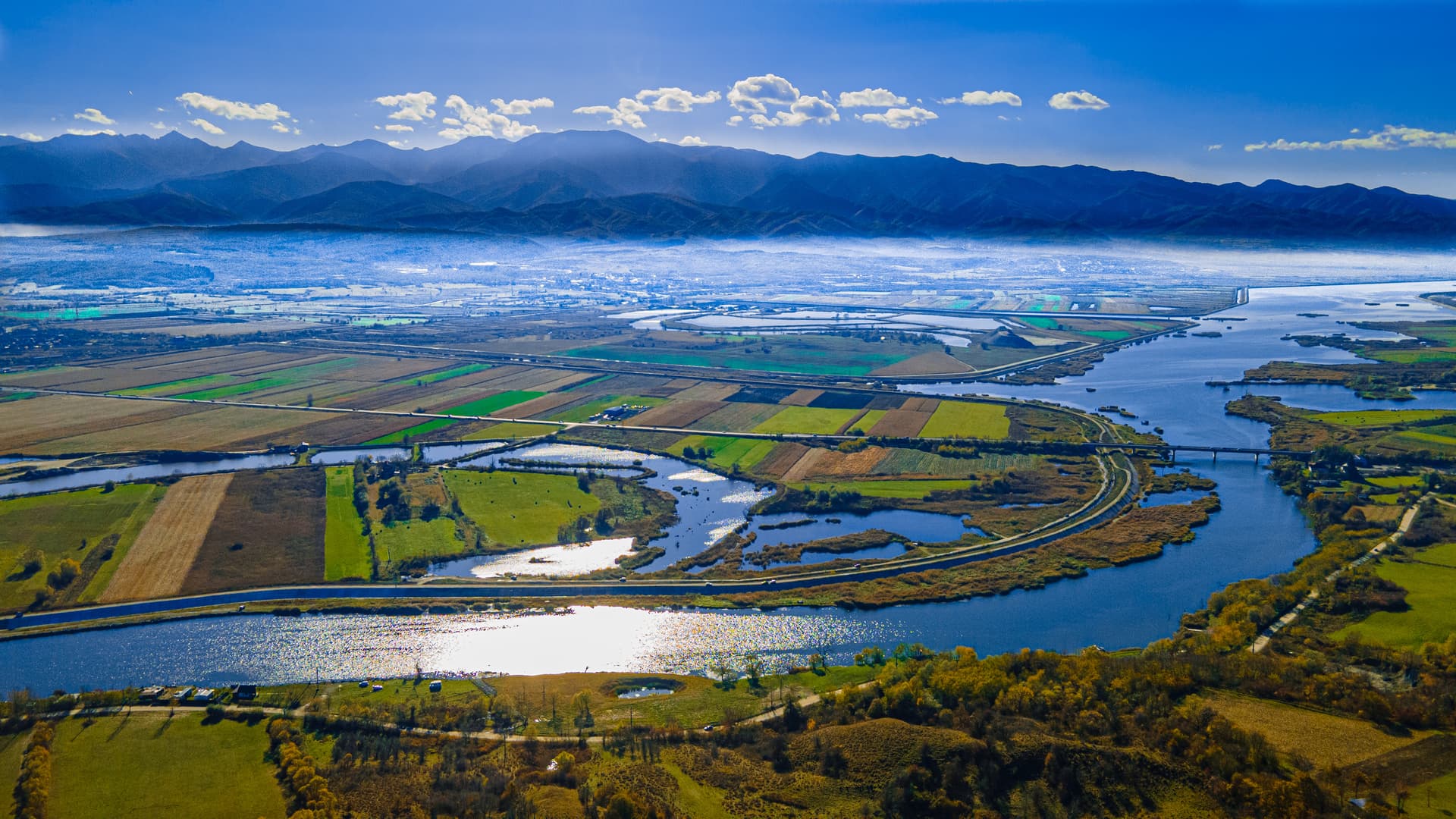 Aerial view of Olt River valley with patchwork fields, water, and Fagaras Mountains in Romania.